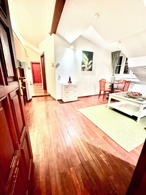 Une pièce spacieuse avec un plancher en bois, un coin repas comprenant une table et des chaises, une commode blanche et une porte rouge.