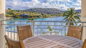 Un balcón con mesa y sillas de madera, con vista a una zona costera de aguas azules cristalinas, palmeras y un paisaje montañoso al fondo.