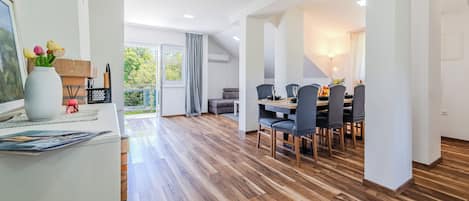 A spacious room with a wooden floor, a dining area with chairs and a table, and a large window with curtains.