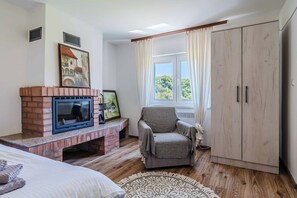 Une chambre avec une cheminée en briques, un fauteuil, une armoire et une fenêtre avec des rideaux.
