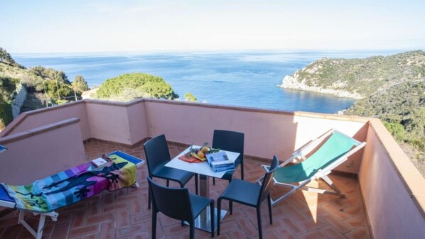 Een terras met een tafel, stoelen, een loungestoel en uitzicht op zee en heuvels.