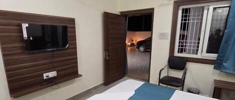 A bedroom with a bed, a ceiling fan, a TV mounted on the wall, and a window with a view of the outside.