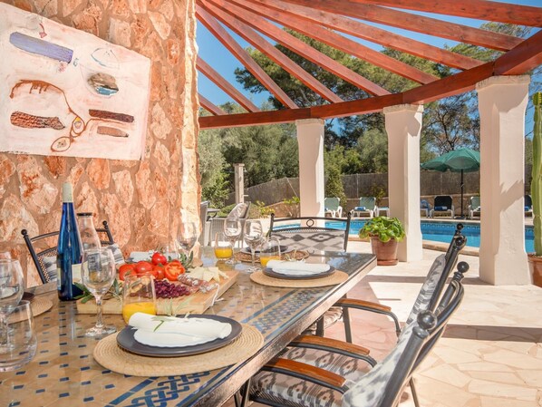Ein Essbereich im Freien mit einer Steinmauer, einer Pergola und einem Pool im Hintergrund.
