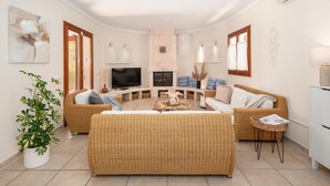 A living room with a fireplace, a television, wicker furniture, and a potted plant. - Villa Sanchez – Comfort and tranquility by the sea. (Santanyi)