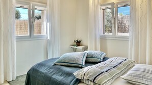 Ein Schlafzimmer mit zwei Fenstern, einem Nachttisch mit Pflanze und einem Bett mit vielen Kissen.