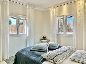 A bedroom with two windows, a bedside table with a plant, and a bed with multiple pillows. - Casa Guapita – Villa with private pool and sea views for 6 guests (Santanyi)