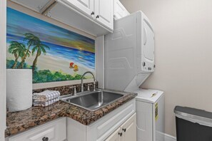A kitchen with a granite countertop, a stainless steel sink, a painting of a beach scene, and a white refrigerator. - The Casita at Salt Therapy | King Bed | Parking (Santa Rosa Beach)