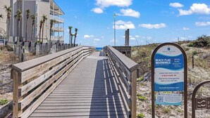 Ein hölzerner Steg führt zum Strand, mit einem Schild, das den Zugang zum Strand und Notfallkontaktdaten anzeigt.