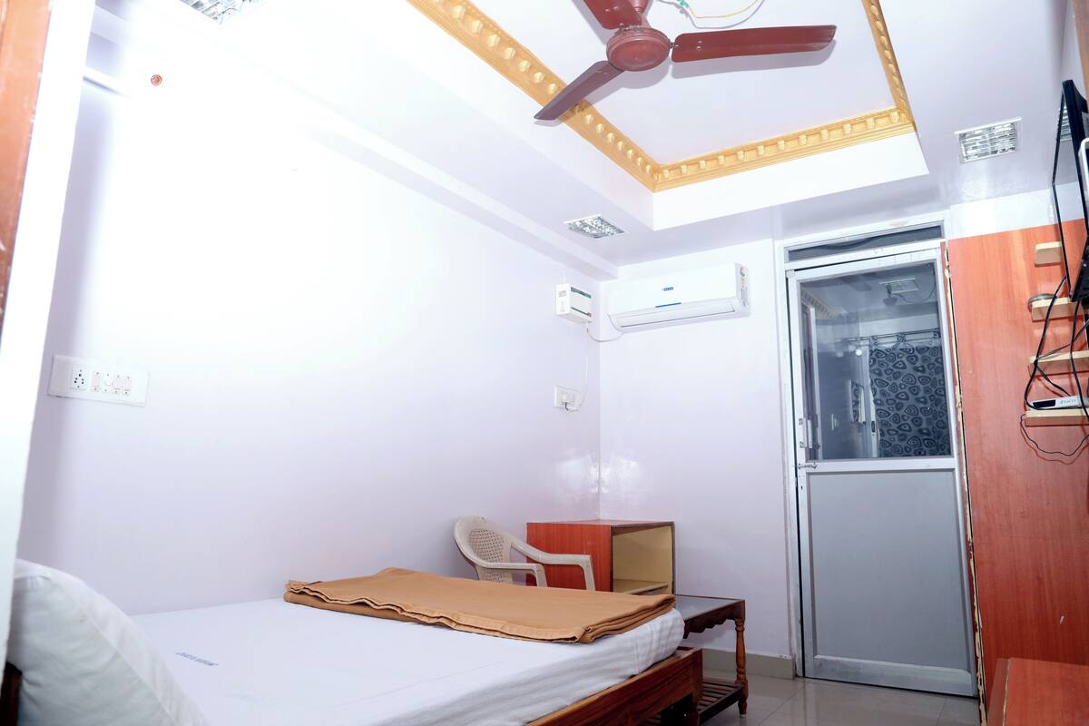 Une petite pièce bien éclairée, avec un lit, un bureau, une chaise et un ventilateur de plafond.