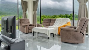 A modern living room with a large flat-screen TV, reclining sofas, a coffee table, and a view of a mountainous landscape through large windows. - Parvata Vista By Soumil's Stays (Karjat)
