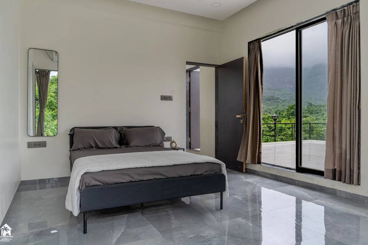 A modern bedroom with a large bed, a mirror, and a view of lush greenery through a sliding glass door.