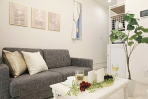 Sala de estar com sofá cinza, mesa de centro branca com uma taça de vinho, uvas e velas. Há três quadros na parede e uma planta à direita.