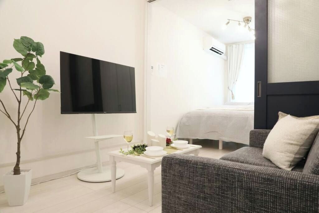 Ein modernes Hotelzimmer mit einem Flachbildfernseher, einem Esstisch mit weißer Tischdecke, einem grauen Sofa und einer Topfpflanze.