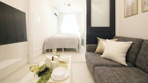 Una moderna camera d'albergo con un divano grigio, una televisione a schermo piatto, una zona pranzo con tavolo apparecchiato per due persone e un letto con copripiumino bianco.