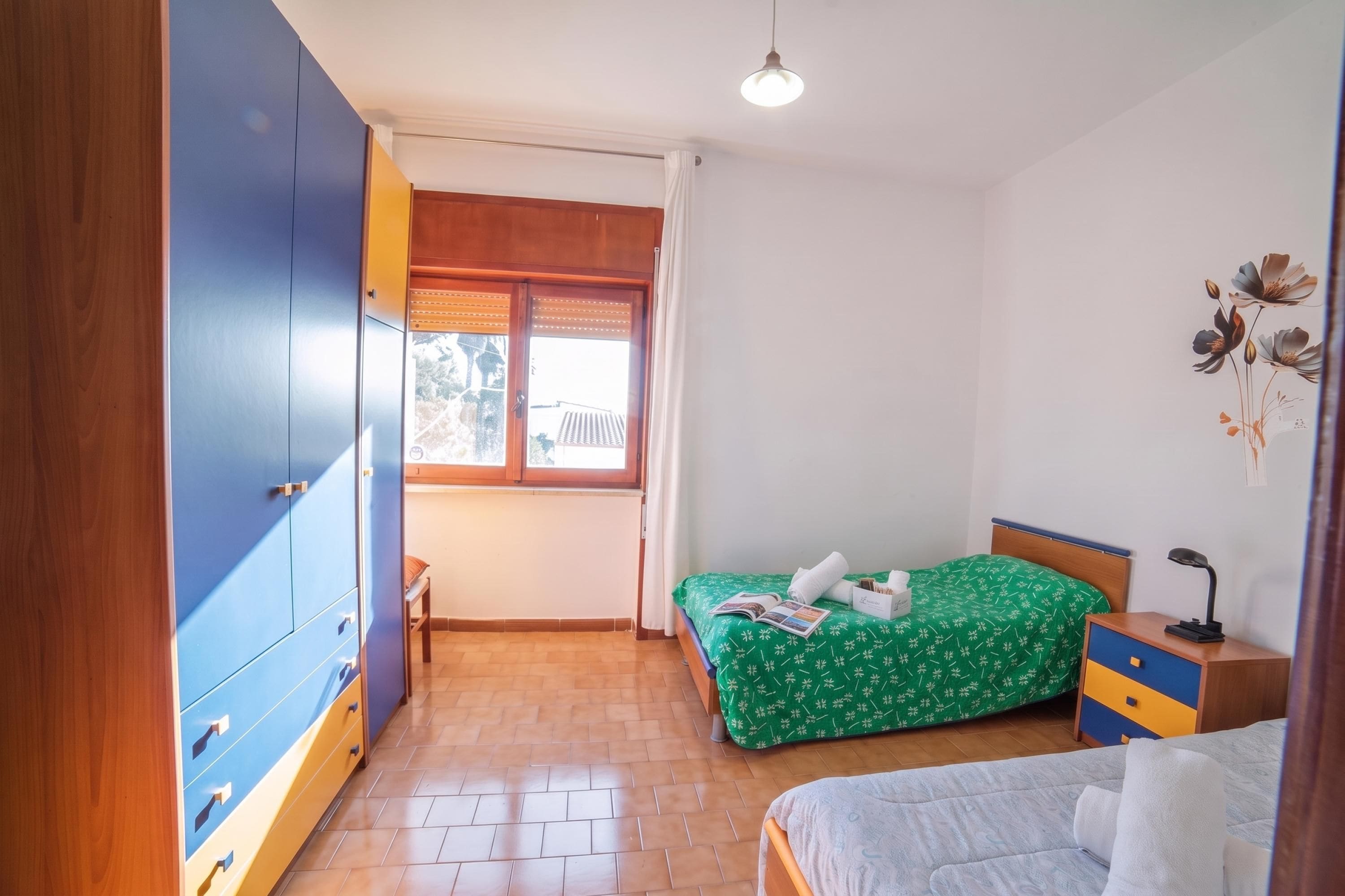 Une chambre à coucher avec un lit, une table de chevet, une lampe, une armoire et une fenêtre avec des rideaux.