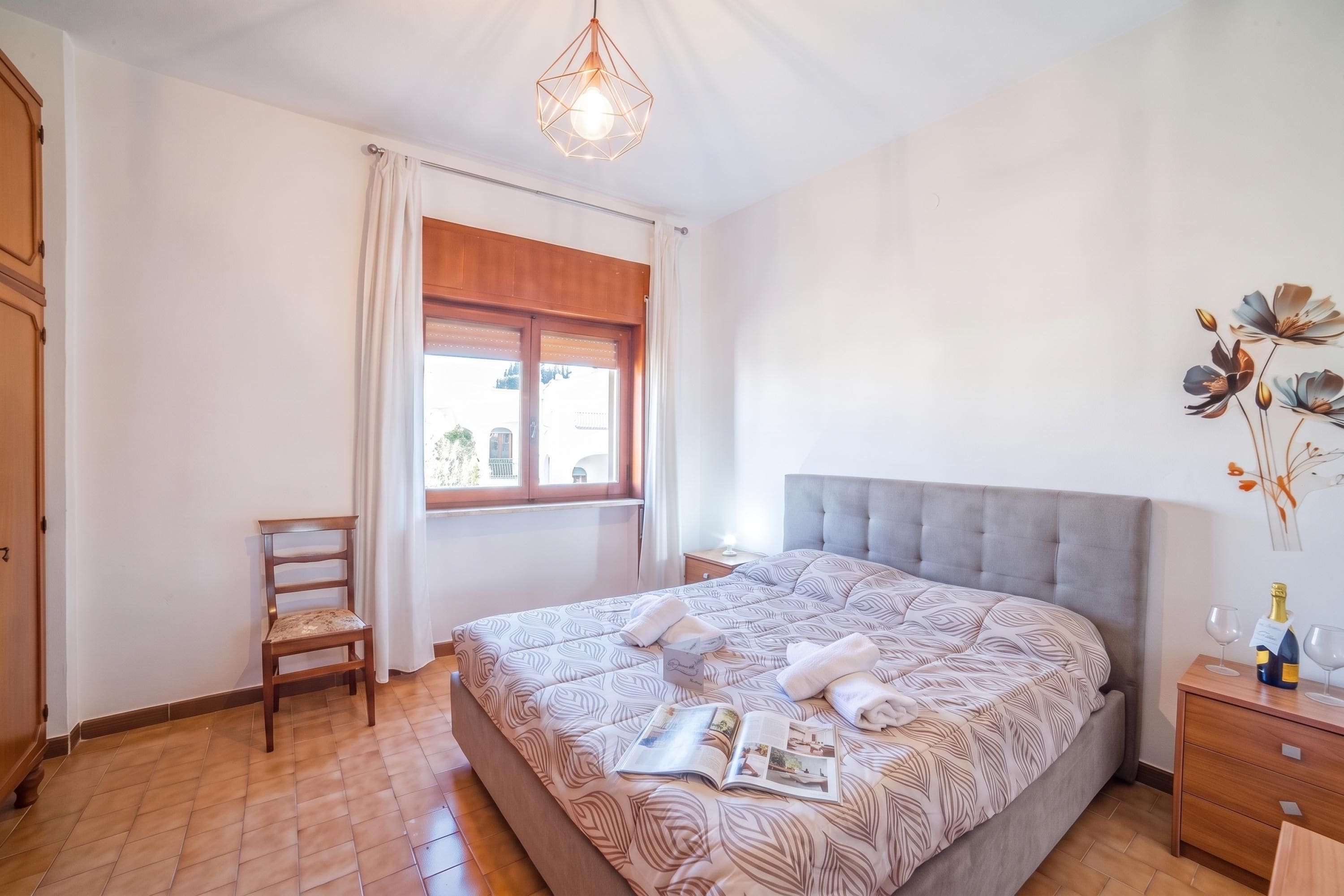 Un dormitorio con una cama grande, una silla de madera, una mesita de noche y una ventana con cortinas.