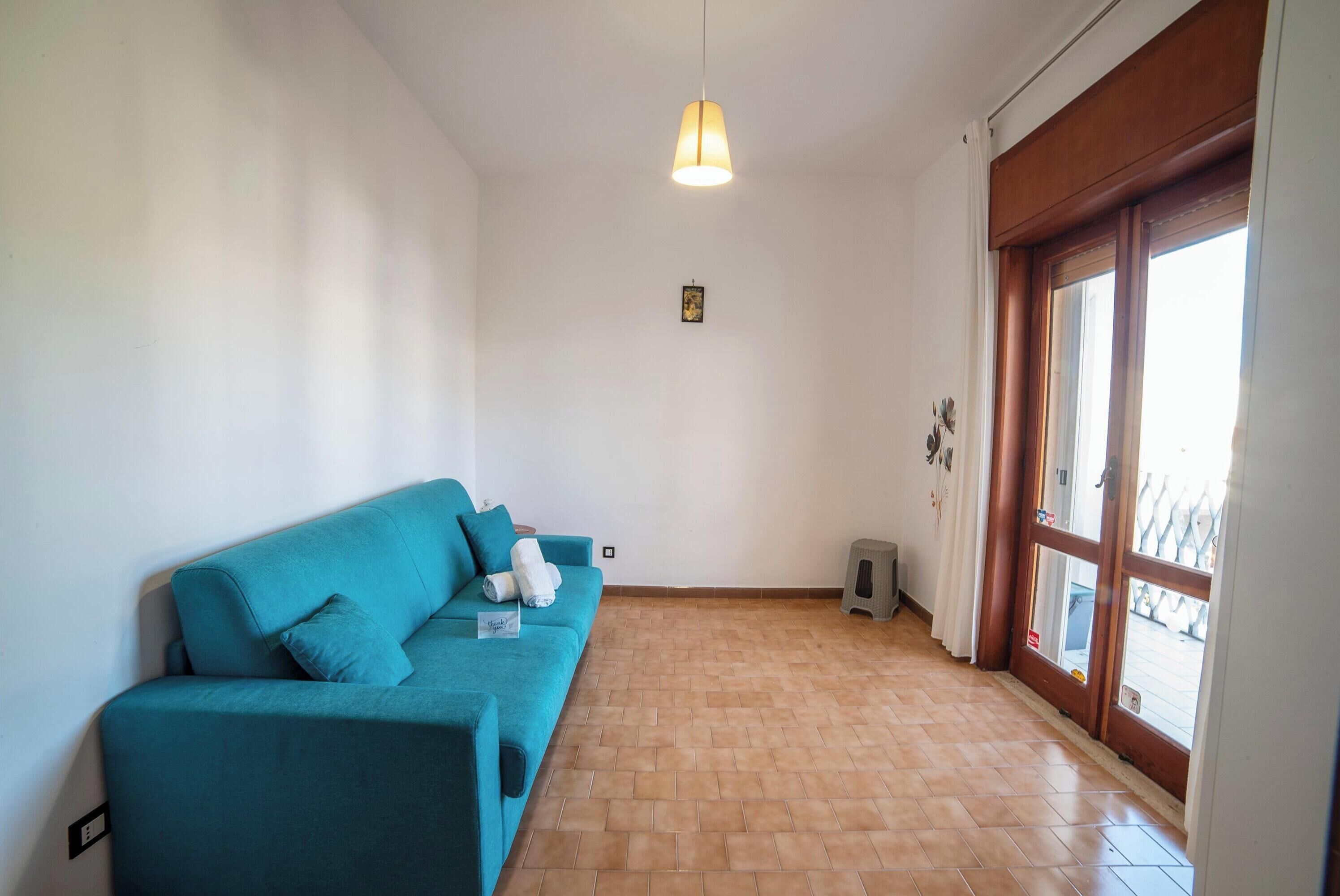 Una habitación con un sofá verde azulado, una lámpara colgante, una puerta de madera y un piso de baldosas.