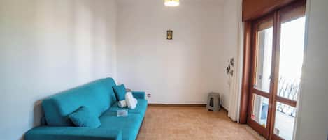 Una habitación con un sofá verde azulado, una lámpara colgante, una puerta de madera y un piso de baldosas.