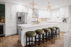 A modern kitchen with white cabinets, a stainless steel refrigerator, a large island with stools, and pendant lighting. - Elevated Old Town Scottsdale Pool| Spa| Sauna|Gym (Scottsdale)