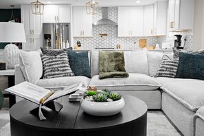 A modern living room with a sectional sofa, a coffee table with a succulent centerpiece, and a kitchen in the background with white cabinets and a hexagonal backsplash. - Elevated Old Town Scottsdale Pool| Spa| Sauna|Gym (Scottsdale)