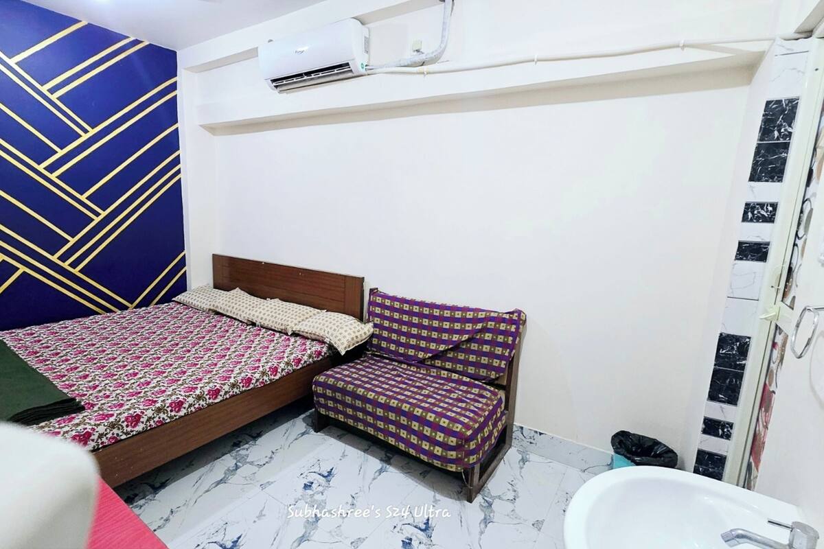 A small, compact bedroom with a bed, a bedside table, a chair, and a bathroom with a sink and toilet.