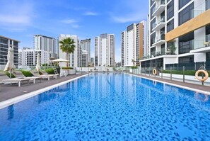 Ein Swimmingpool mit Liegestühlen und Sonnenschirmen, umgeben von modernen Hochhäusern.