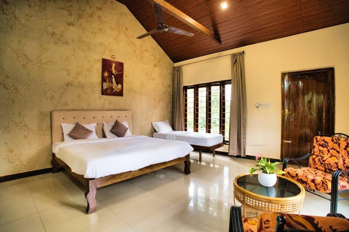 A room with two beds, a ceiling fan, a window with curtains, a door, and a chair with a patterned cushion.