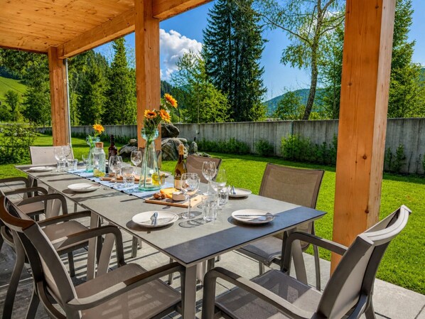 Ein Essbereich im Freien mit einer Holz-Pergola, einem Tisch für sechs Personen mit Stühlen und Blick auf einen Rasen und Bäume.