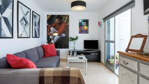 Een moderne woonkamer met een grijze bank, rode kussens, een salontafel met een vaas, een flatscreen televisie en kunstwerken aan de muur.