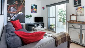 Een moderne woonkamer met een grijze bank, rode kussens, een flatscreen televisie, een klein bijzettafeltje en een boekenkast.