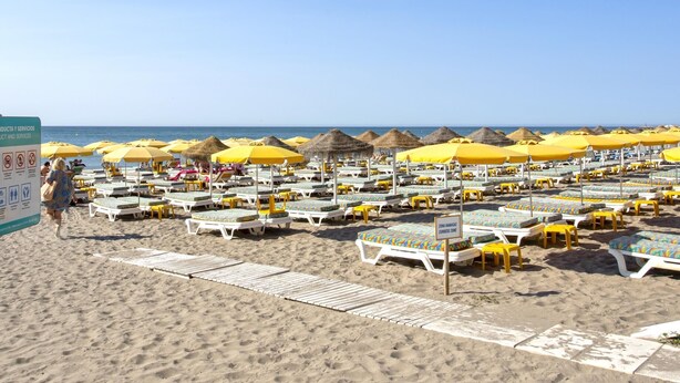 Een strand met rijen ligbedden en gele parasols, een bordje dat een zonnebadzone aangeeft, en een helderblauwe lucht.