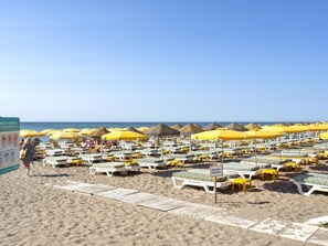 Une plage bordée de rangées de transats et de parasols jaunes, un panneau indiquant une zone réservée aux bains de soleil, et un ciel d’un bleu limpide.
