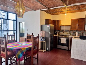 Une cuisine avec des meubles en bois, un réfrigérateur en acier inoxydable, une cuisinière à gaz et une table à manger recouverte d’une nappe colorée.