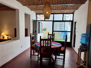 A dining area with a round table covered by a colorful tablecloth, wooden chairs, a hanging light fixture, and a view through large windows. - Central Ajijic 3-Bedroom House, Near Everything! (Ajijic)