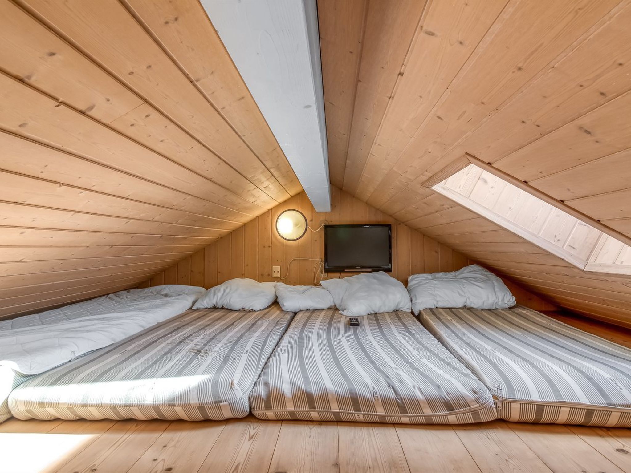 Un dormitorio acogedor en el ático con paredes de madera y techo inclinado. Cuenta con una cama grande con ropa de cama a rayas y dos almohadas, un televisor de pantalla plana montado en la pared, y un tragaluz que proporciona luz natural.