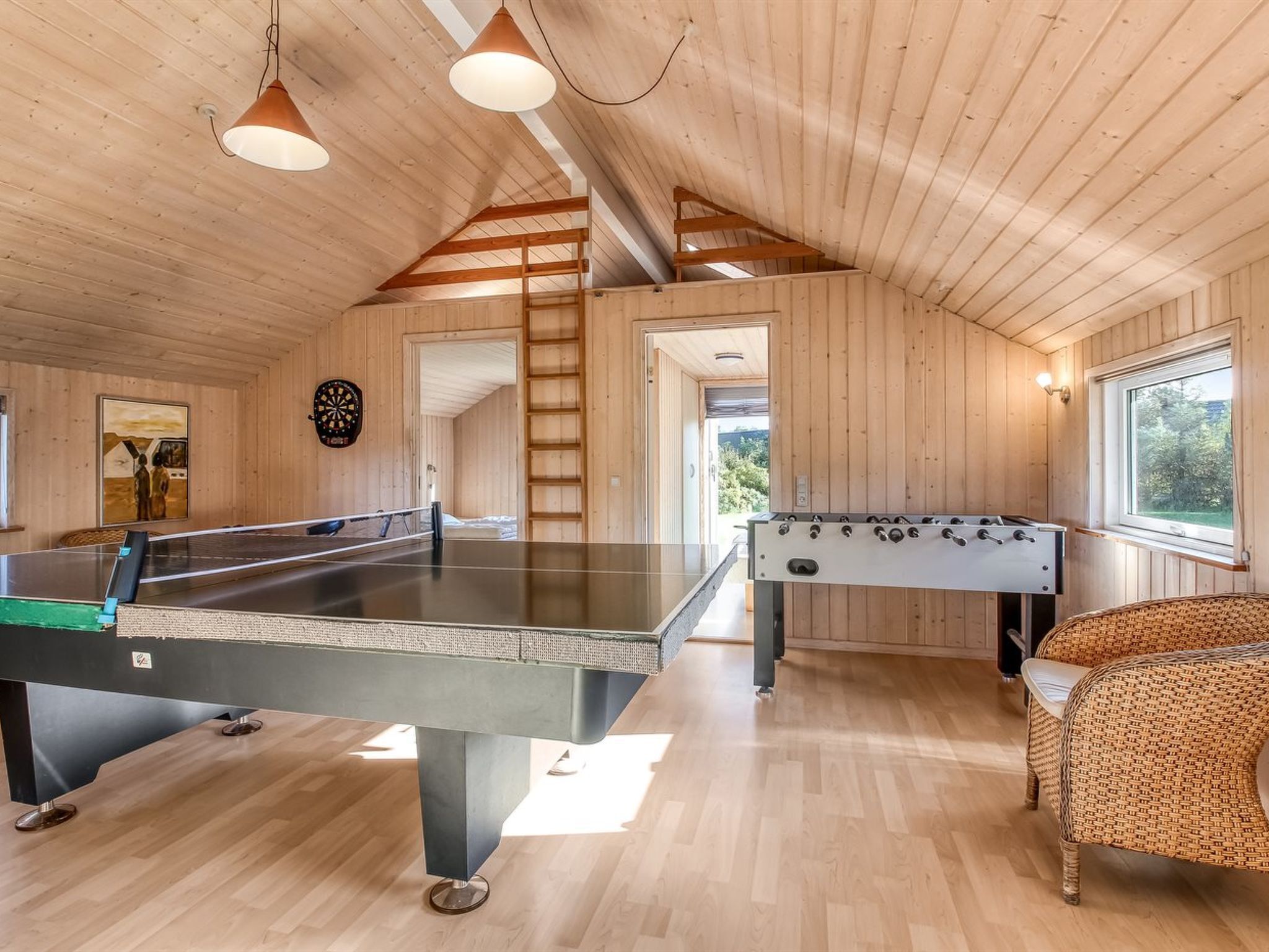 A wooden-floored room with a foosball table, ping pong table, and dartboard.