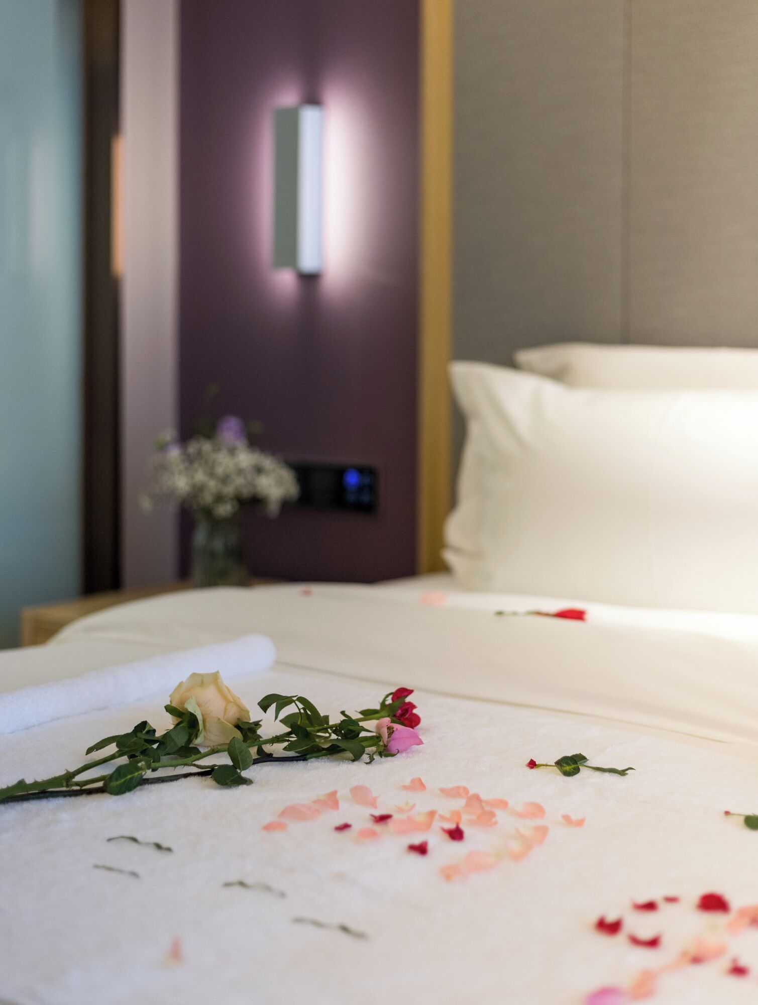A hotel room with a neatly made bed, a vase with flowers, and a mirror on the wall.
