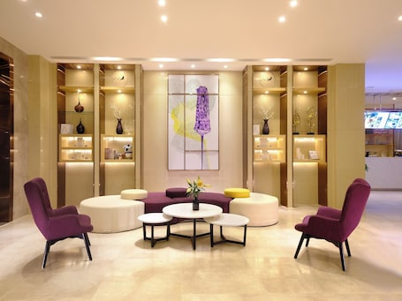 A modern lobby with a central seating area, including purple chairs and a sofa, surrounded by shelves with decorative items and a large abstract painting.