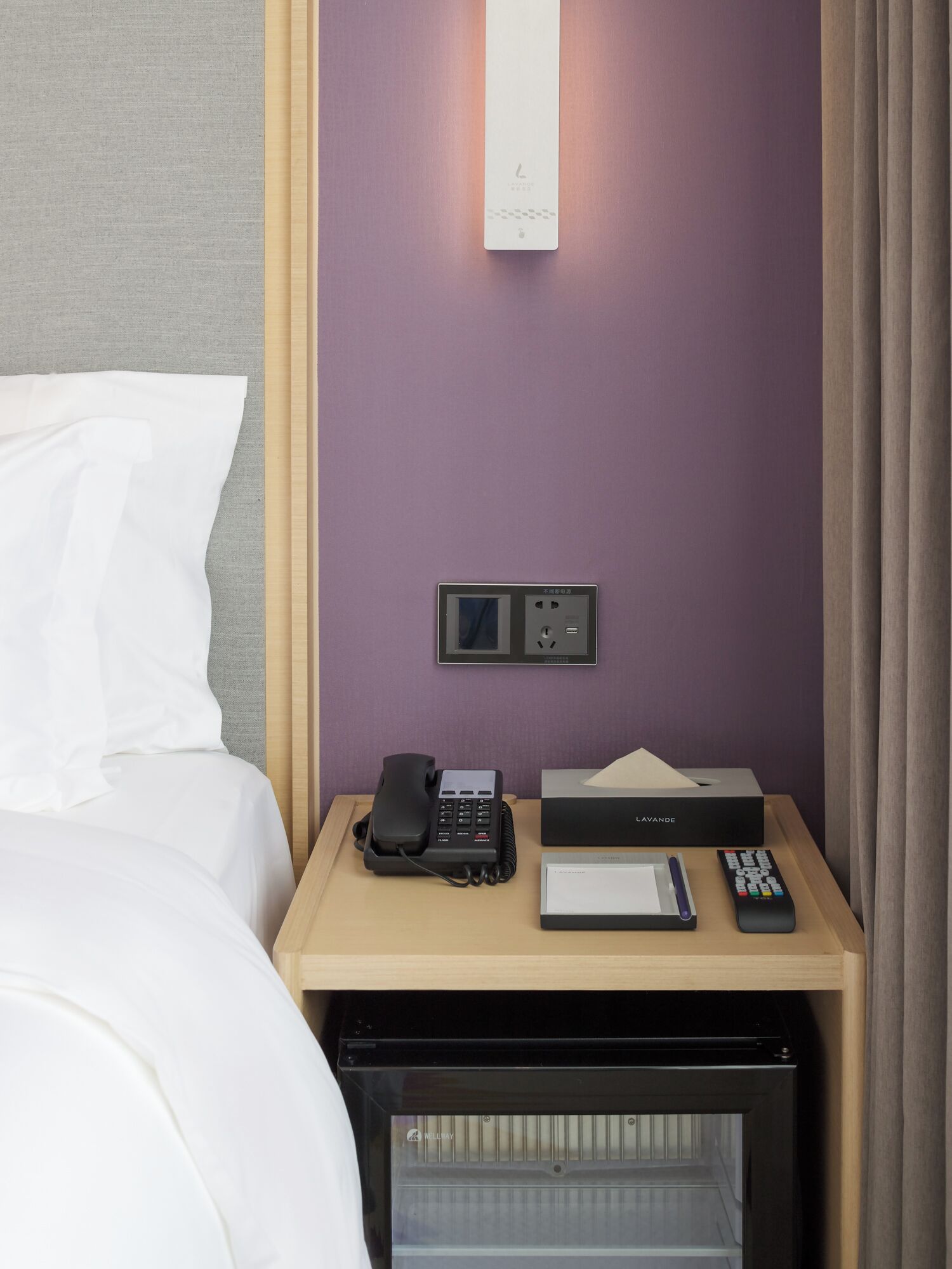 A hotel room with a bed, a bedside table with a phone, a remote control, a tissue box, and a minibar.