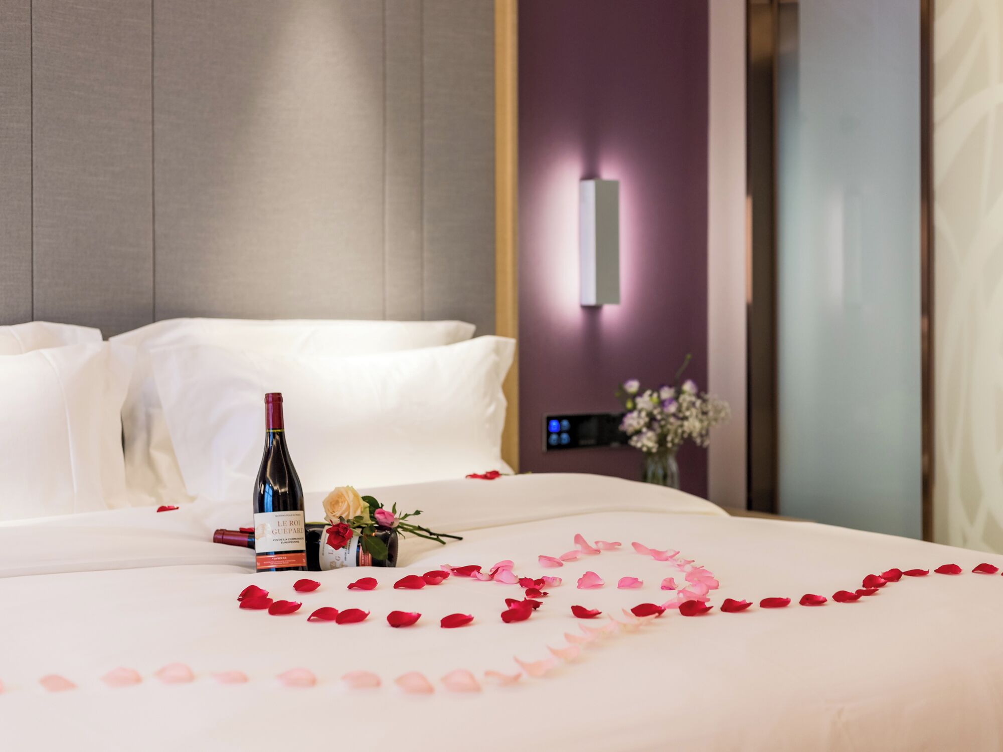 Ein Hotelzimmer mit einem ordentlich bezogenem Bett, einer Weinflasche und Rosenblüten, die auf dem Bett drapiert sind.