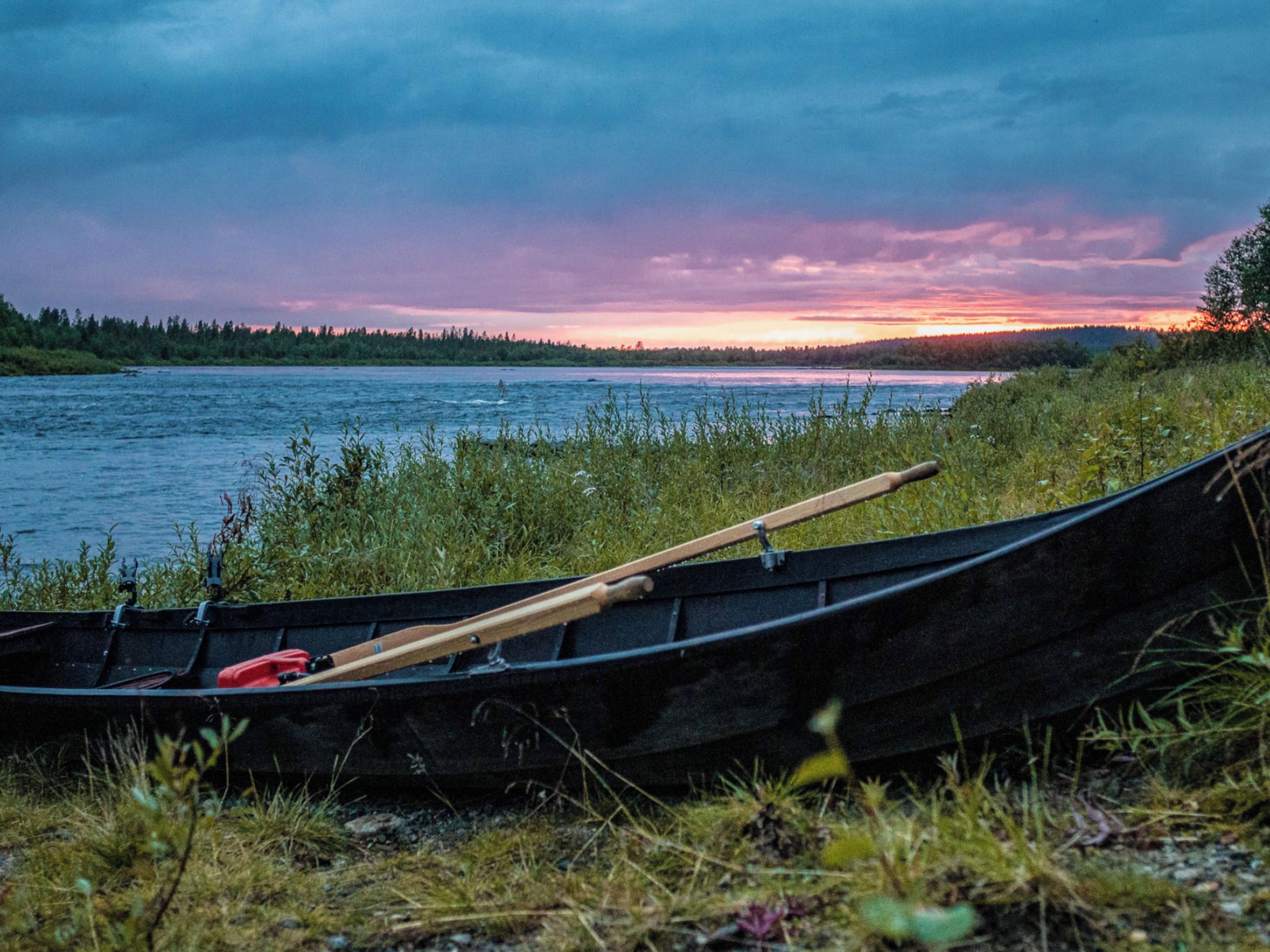 A canoe on the shore with a paddle and a red object, surrounded by grass and a body of water.