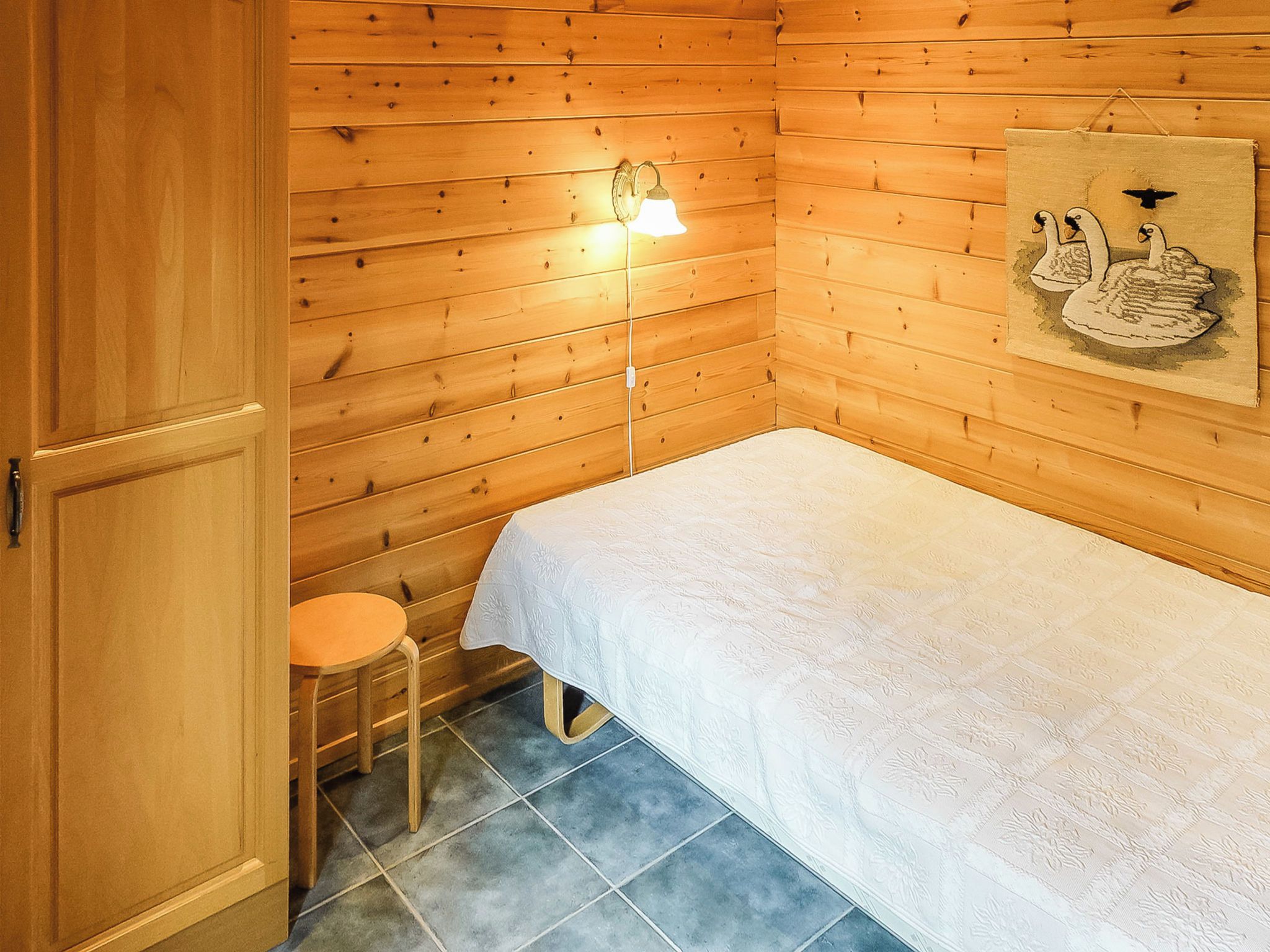 Een kleine, gezellige kamer met houten wanden, een enkel bed met een witte spreideken, een klein rond tafeltje en een wandlamp.