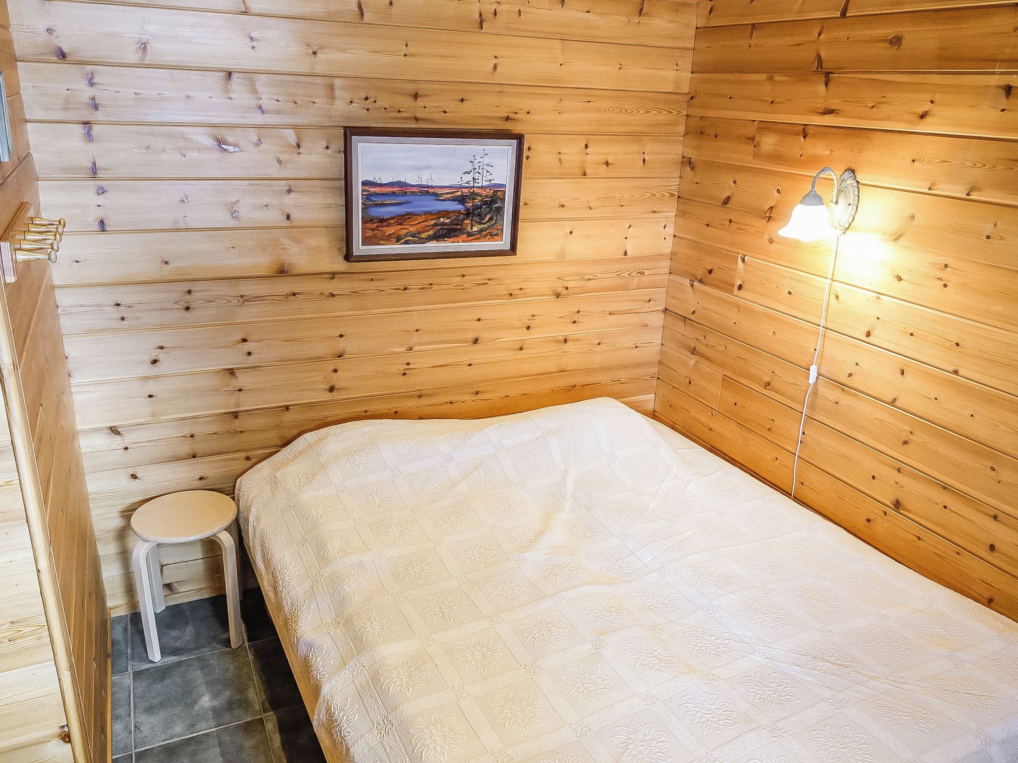 Una habitación de cabaña de madera con una cama, un taburete pequeño y un cuadro en la pared.