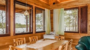 A wooden cabin interior with a dining table, chairs, and large windows overlooking a forested area.