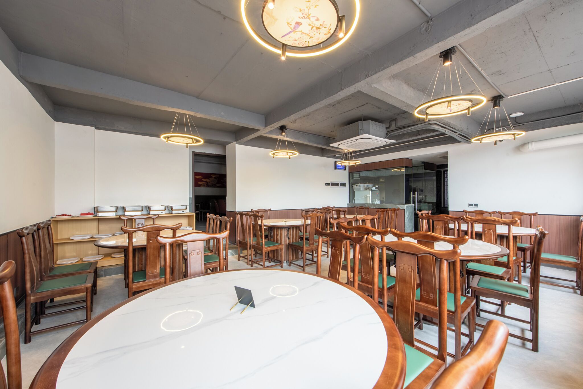 A restaurant with round tables and wooden chairs, overhead lighting, and a visible kitchen area.