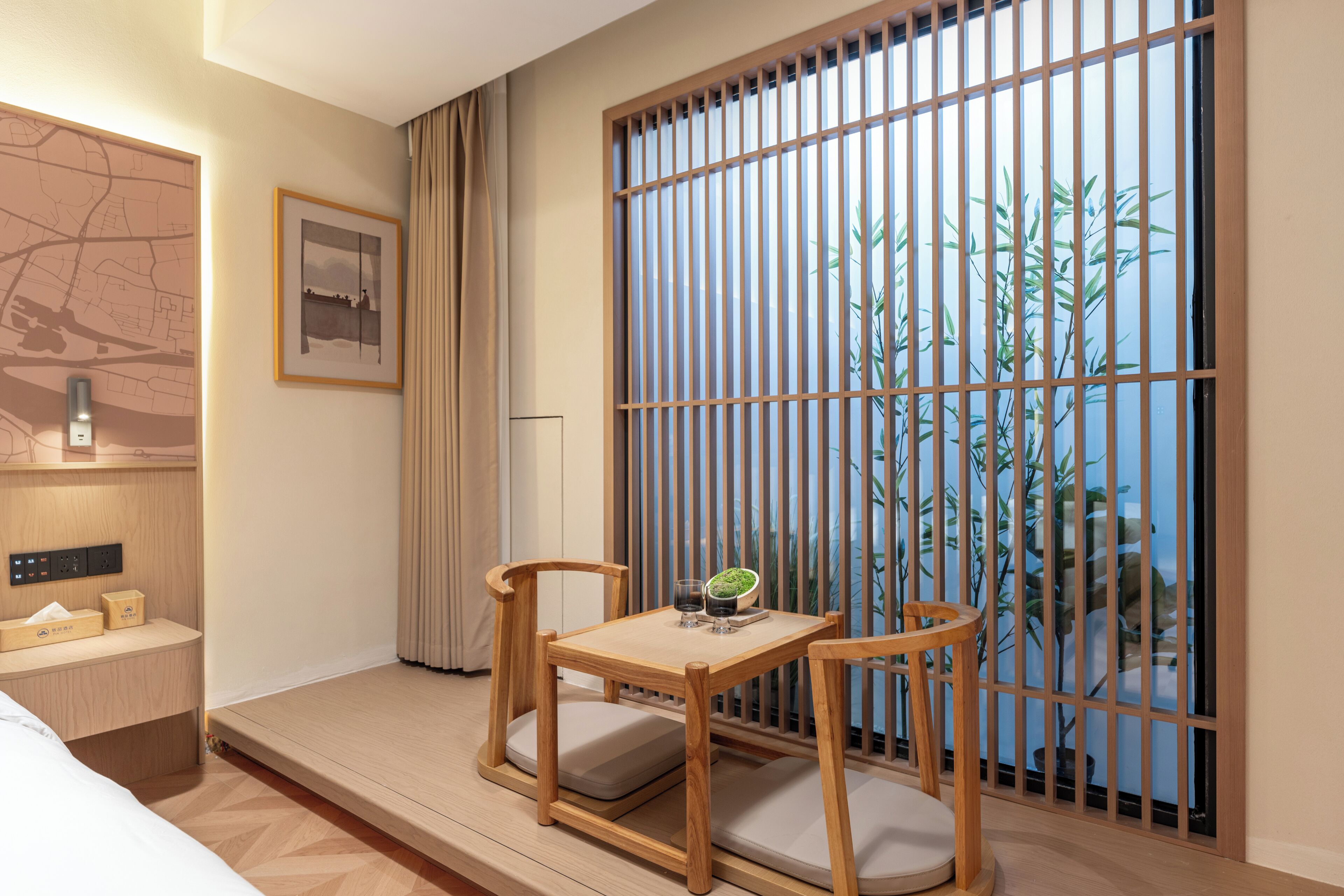 Une chambre d’hôtel avec un lit, une petite table avec des chaises, une carte accrochée au mur et une grande fenêtre donnant sur des plantes de bambou.