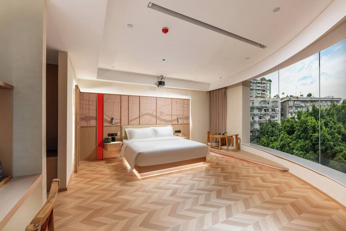 Ein modernes Schlafzimmer mit einem großen Bett, einem Nachttisch, einem kleinen Tisch und Blick auf eine Stadtlandschaft durch ein großes Fenster.