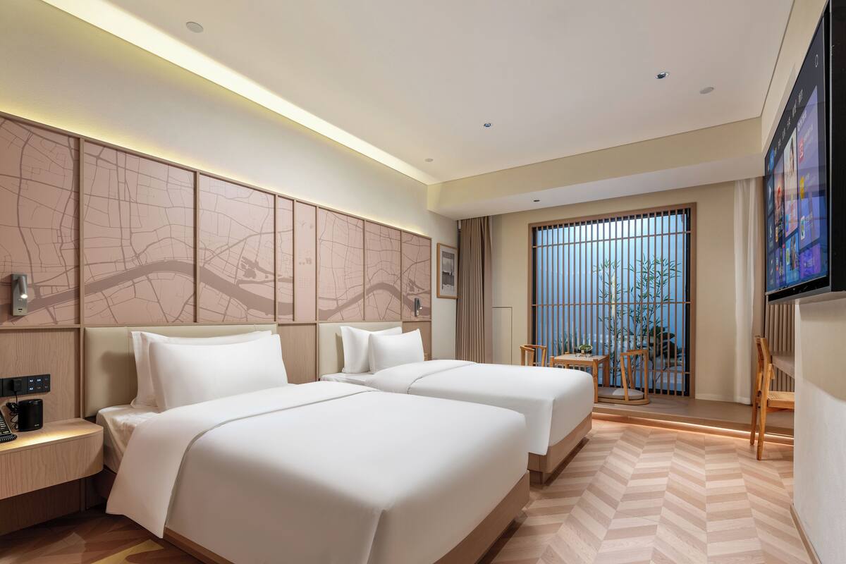 Ein Hotelzimmer mit zwei Betten, einem Schreibtisch mit Telefon, einem Fernseher und einem großen Fenster mit Blick ins Grüne.