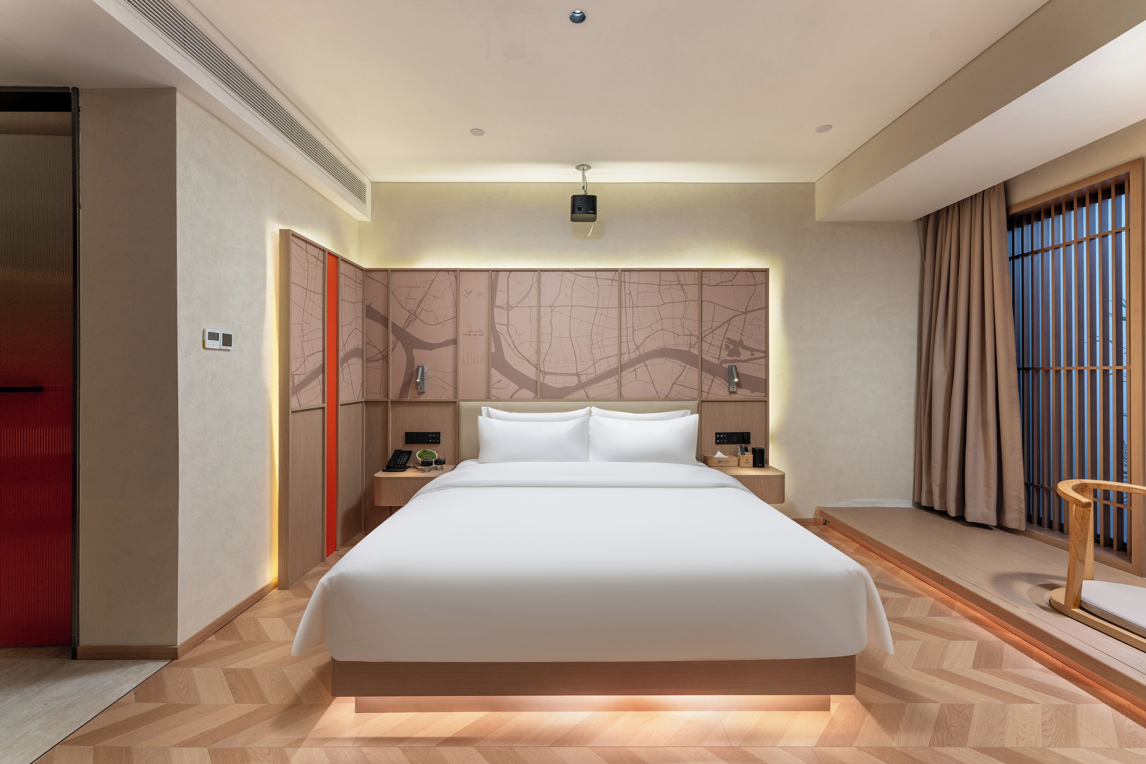 A modern hotel room with a large bed, a wooden floor, a map wall, and a view of the outside through a window with curtains.