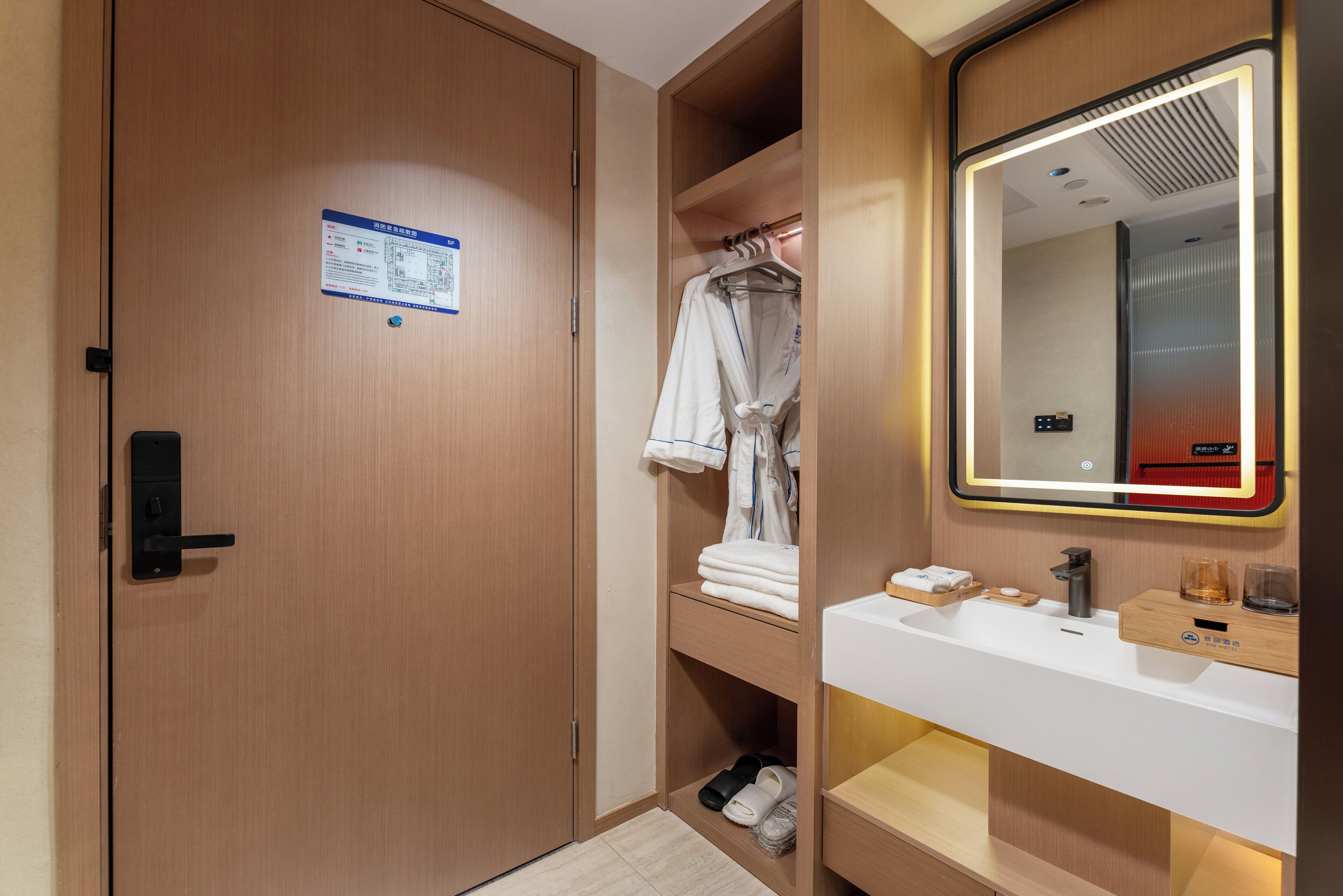 A hotel bathroom with a wooden door, a towel rack with folded towels, a sink with a mirror, and a small shelf with toiletries.